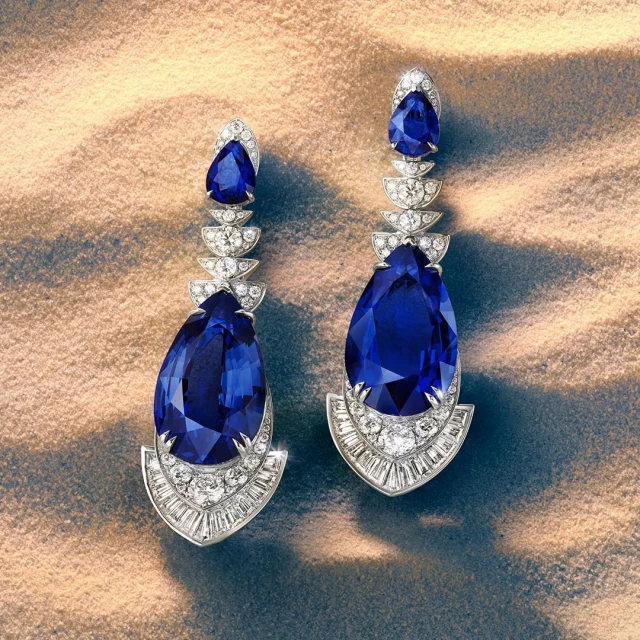 Elegant teardrop earrings with blue gemstones and diamonds on a textured beige surface, captured in a detailed studio shot.