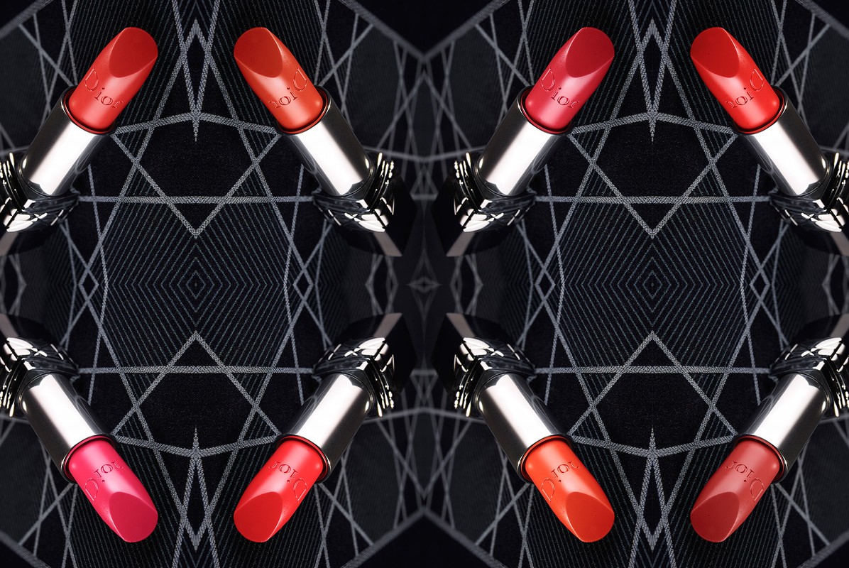 Symmetrical close-up of lipstick tubes in various red shades with silver casings, arranged on a black geometric-patterned backdrop.