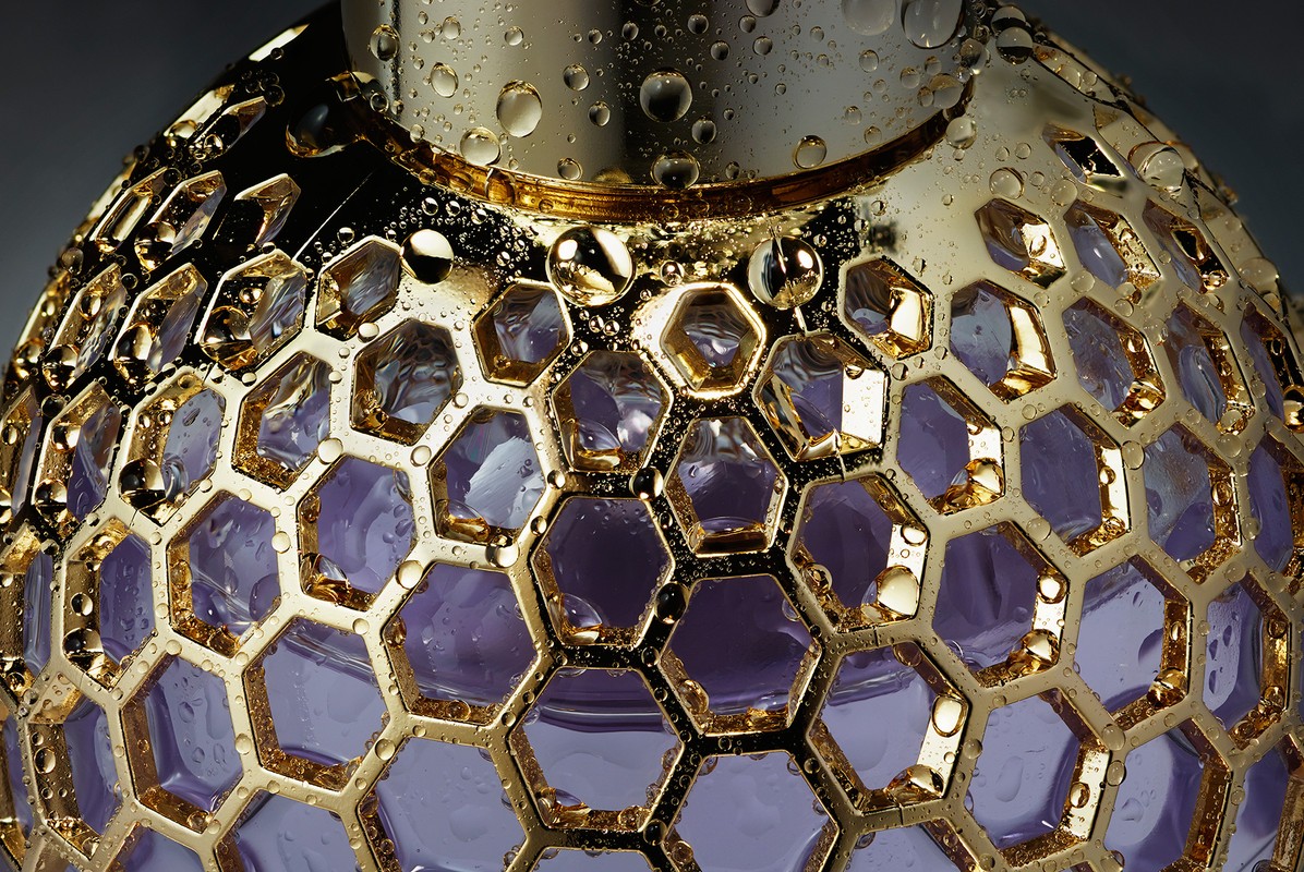 Close-up of a gold perfume bottle with a purple liquid interior and water droplets on its surface, showcasing luxury and detail.