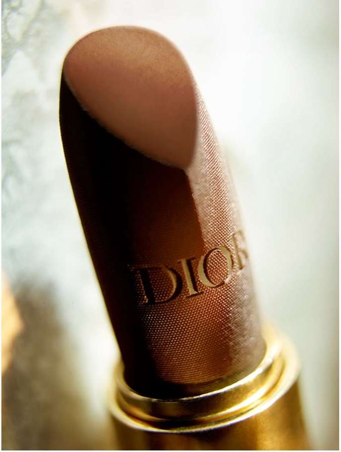 Close-up of a Dior lipstick with embossed lettering in a metallic gold case against a soft textured background.