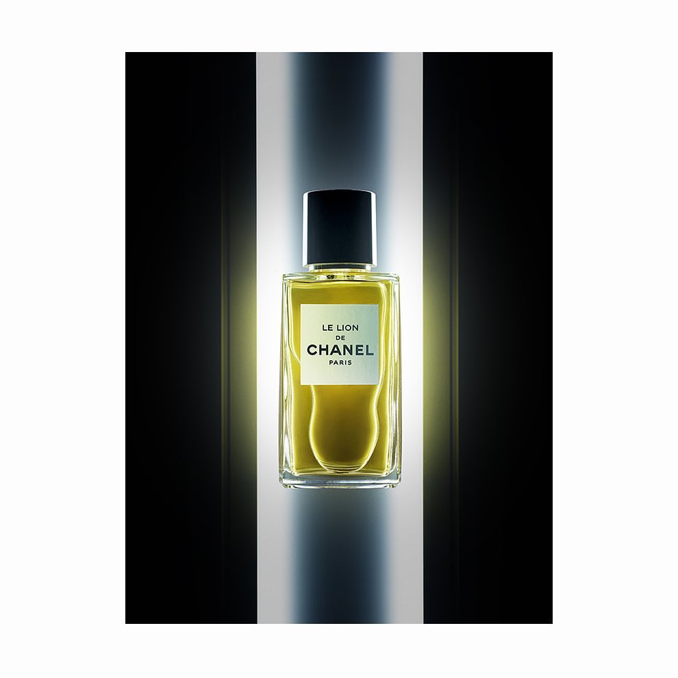 A rectangular glass perfume bottle with a black cap centered against a vertical illuminated column and dark background.
