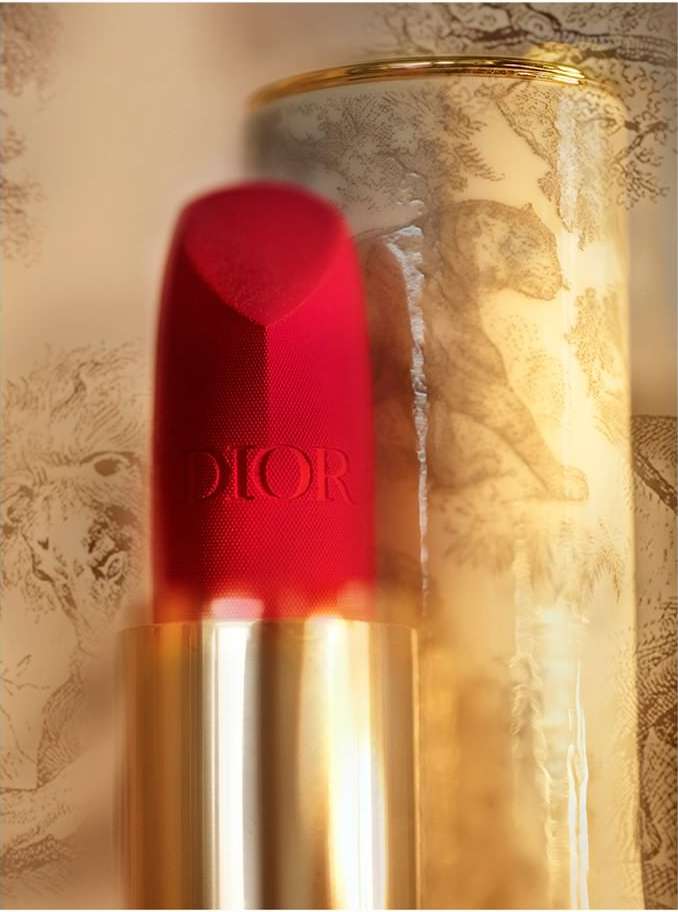 Close-up of red Dior lipstick with a gold base, beside a patterned canister featuring sketched animals. Beauty and cosmetics product shot.