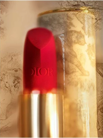 Close-up of red Dior lipstick with a gold base, beside a patterned canister featuring sketched animals. Beauty and cosmetics product shot.