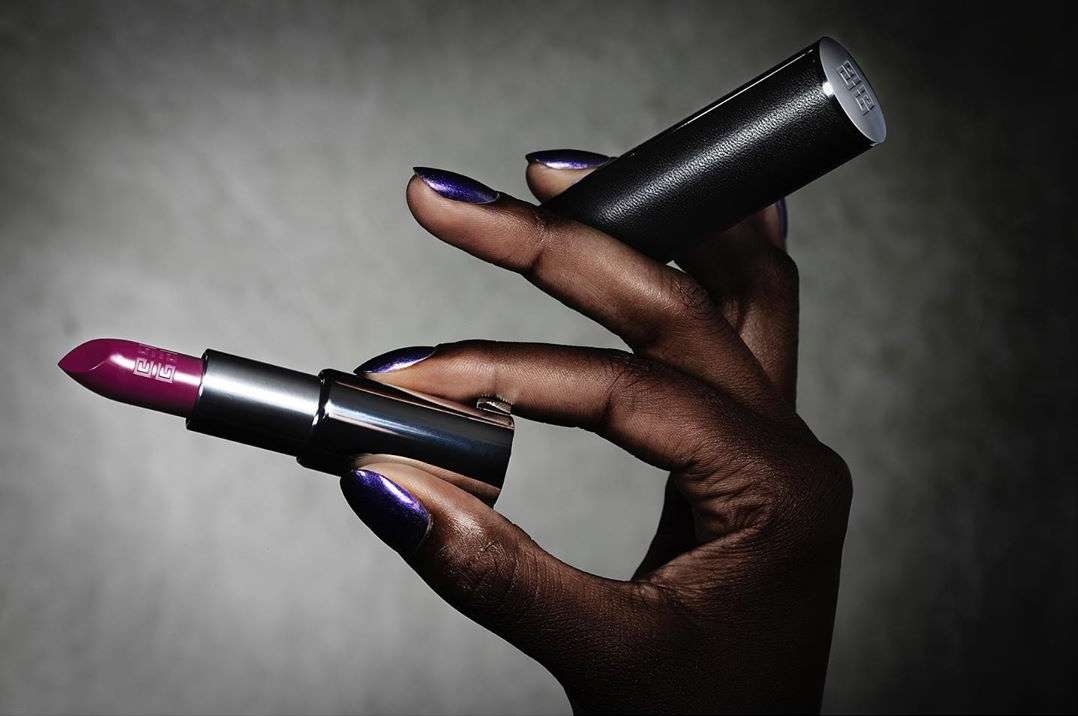 Close-up of a dark-skinned hand holding a tube of deep purple lipstick and its black cap, beauty shot against a gradient background.