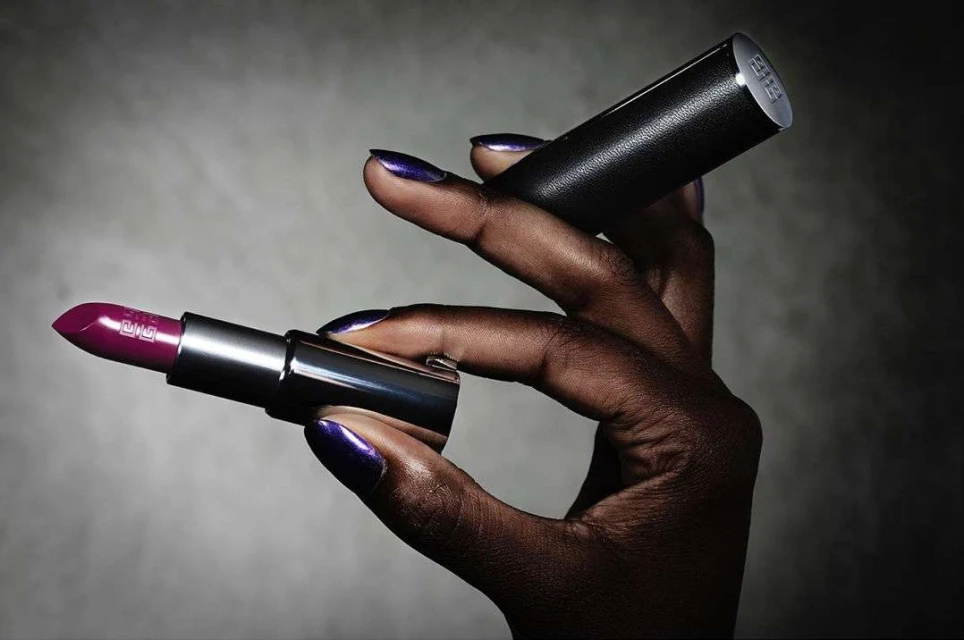 Close-up of a dark-skinned hand holding a tube of deep purple lipstick and its black cap, beauty shot against a gradient background.