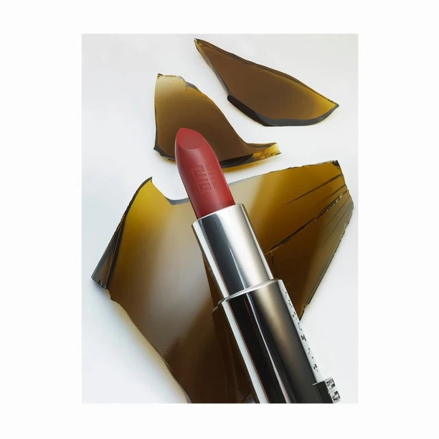 Close-up studio shot of a red lipstick tube surrounded by shards of brown glass, set against a clean white backdrop.