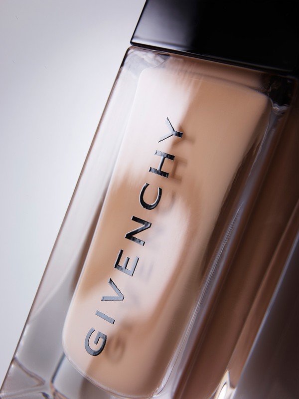 Close-up of a clear glass bottle of Givenchy foundation make-up with a black cap on a white background, lit with studio lighting.