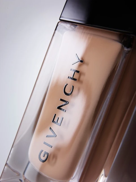 Close-up of a clear glass bottle of Givenchy foundation make-up with a black cap on a white background, lit with studio lighting.