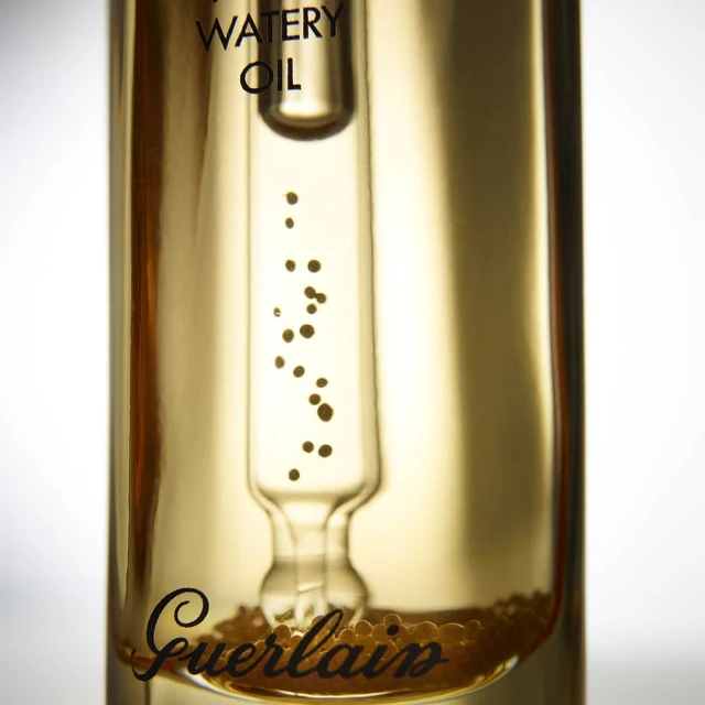 Close-up of Guerlain Abeille Royale Watery Oil skincare product in clear glass bottle with visible oil droplets and product text.