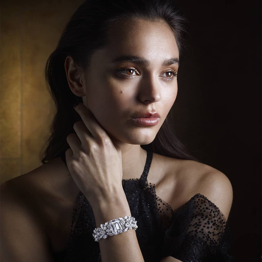 Portrait of a woman with brown hair wearing a black dress and a white jeweled bracelet against a dark gold background.