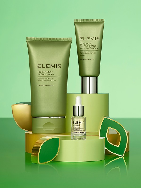 Three Elemis skincare products are displayed on a green surface, including facial wash, jelly exfoliator, and serum, creating a beauty still life.