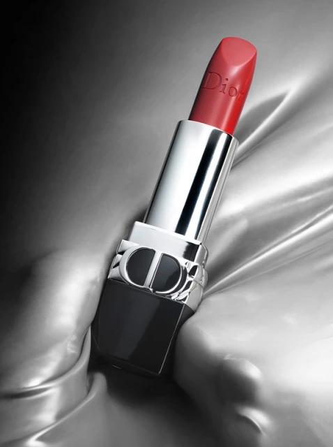 Red lipstick on a shiny, silver fabric background. The lipstick has a black and silver tube with the Dior logo.
