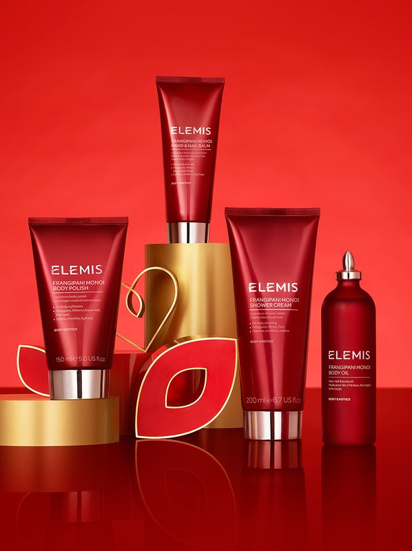 Elevated view of Elemis skincare products in red and gold packaging arranged on a red reflective surface against a red backdrop.