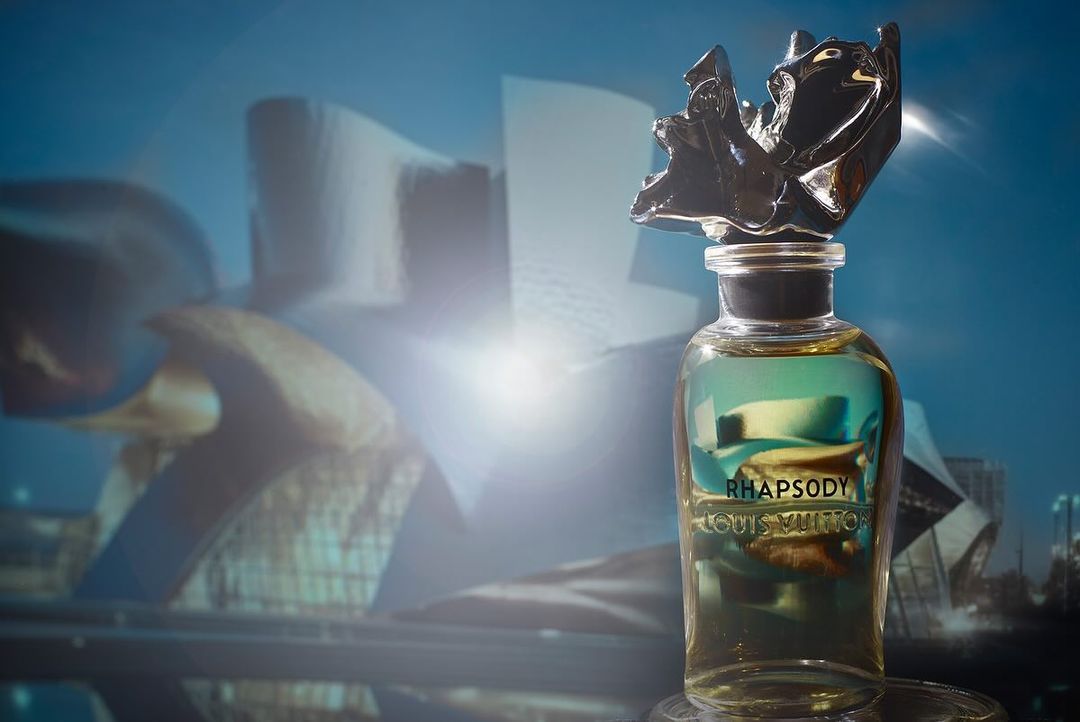 Louis Vuitton perfume bottle of Rhapsody in a glass container with an artistic black topper, set against a blurred architectural backdrop.