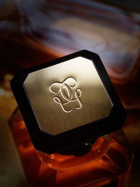 Close-up shot of the lid of a Guerlain fragrance bottle with an ornate logo and a gradient background featuring amber, gold, and brown tones.