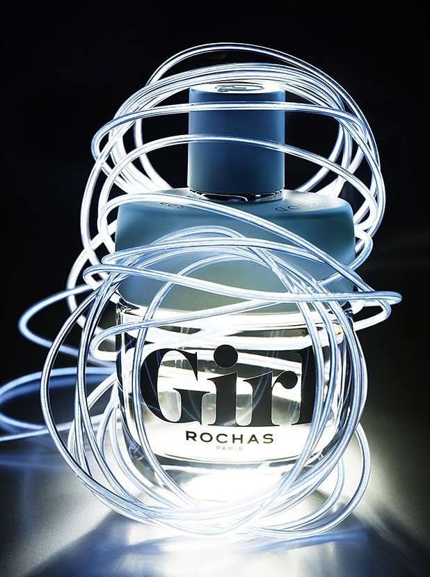 A bottle of Rochas Girl perfume is displayed in a studio setting, wrapped in glowing, flexible white tubing against a dark backdrop.