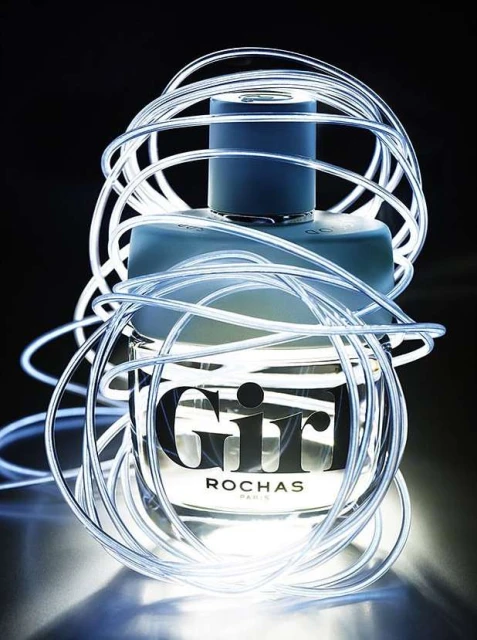 A bottle of Rochas Girl perfume is displayed in a studio setting, wrapped in glowing, flexible white tubing against a dark backdrop.