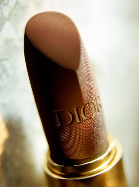 Close-up studio shot of a brown Dior lipstick with golden base, showcasing the brand's embossed logo, on a soft blurred background.