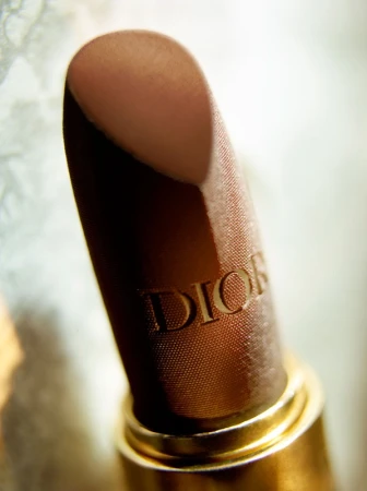 Close-up studio shot of a brown Dior lipstick with golden base, showcasing the brand's embossed logo, on a soft blurred background.