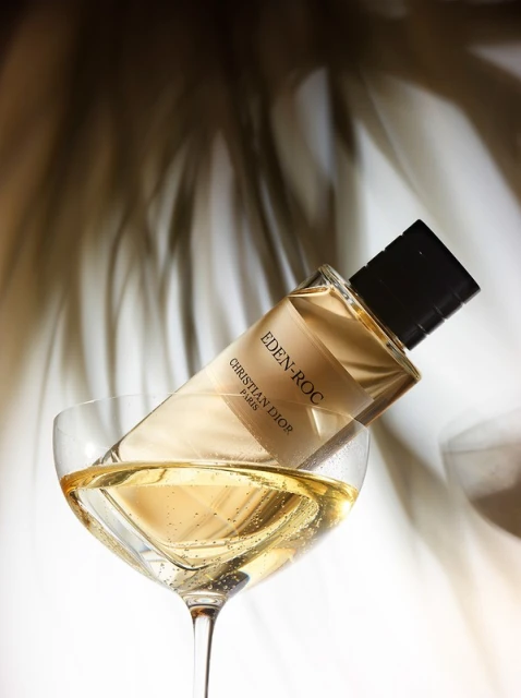 Crystal glass containing a golden perfume bottle of Christian Dior's Eden-Roc, casting a soft shadow on a white background.