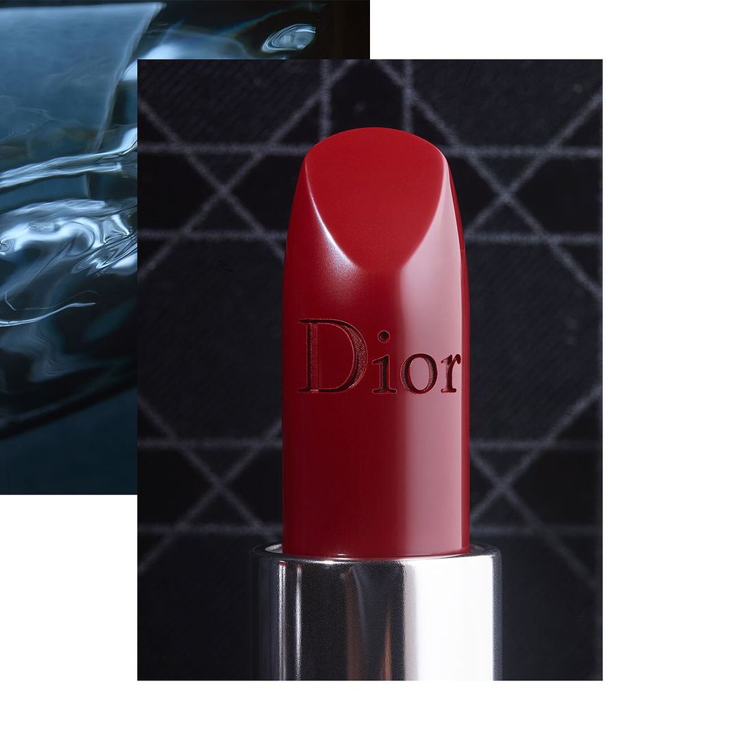 Close-up product shot of a red lipstick with the Dior logo embossed on the side against a dark patterned background.
