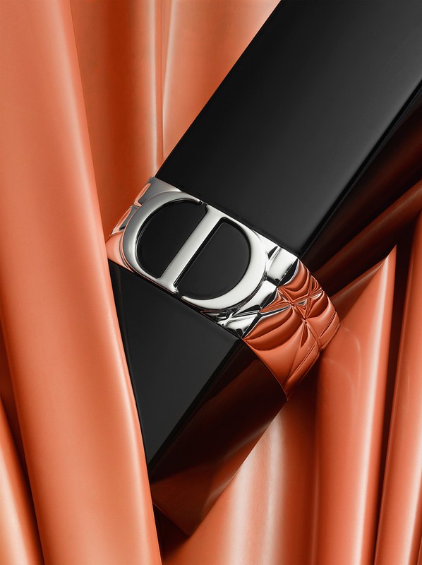 Close-up of a black and silver lipstick tube with a "CD" logo against a peach-colored draped fabric backdrop, beauty, cosmetics.