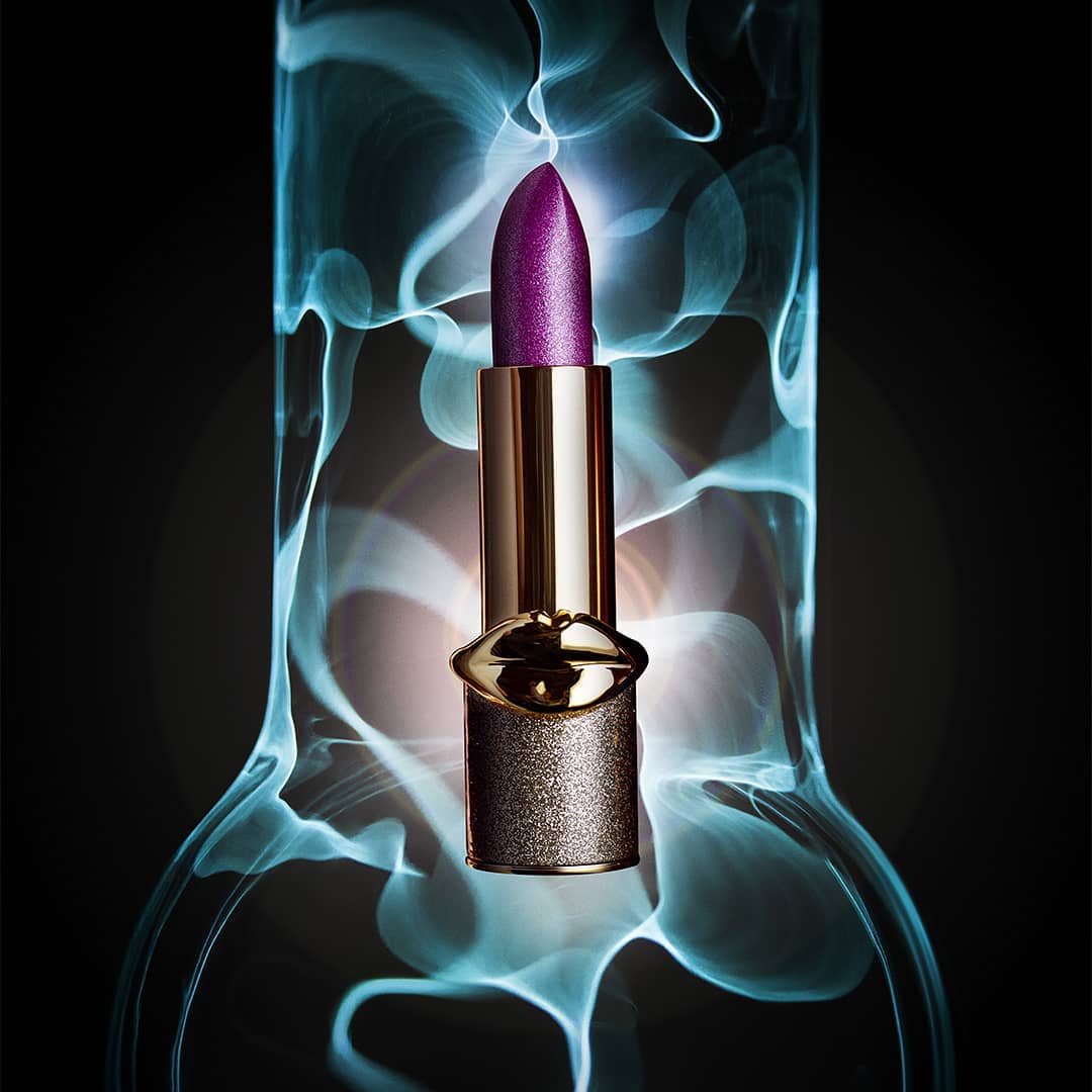 Close-up product shot of shimmering gold lipstick with a metallic purple top against a soft blue glowing background.