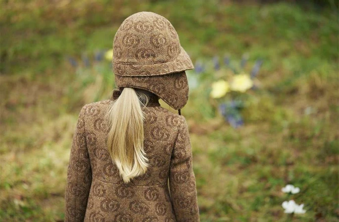 Model seen from behind wearing a patterned brown tweed coat and matching hat with earflaps, outdoors with soft lighting