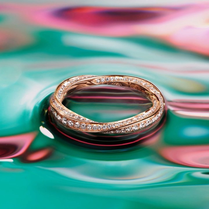 Close-up of a gold intertwined ring with diamond embellishments, showcased on a teal and pink swirling reflective surface.