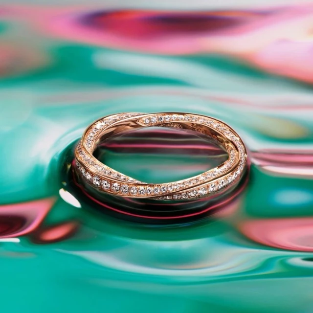 Close-up of a gold intertwined ring with diamond embellishments, showcased on a teal and pink swirling reflective surface.
