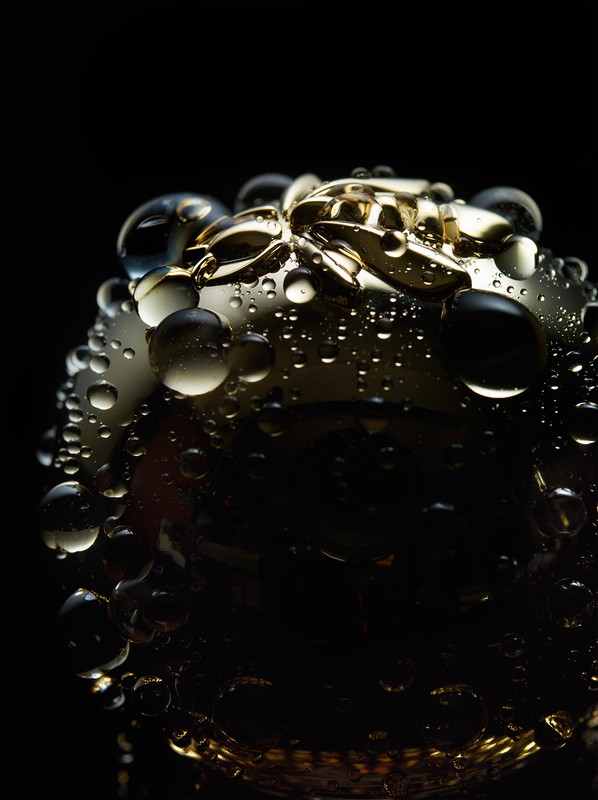 Close-up studio shot of an abstract gold-toned, round object adorned with water droplets against a stark black background.