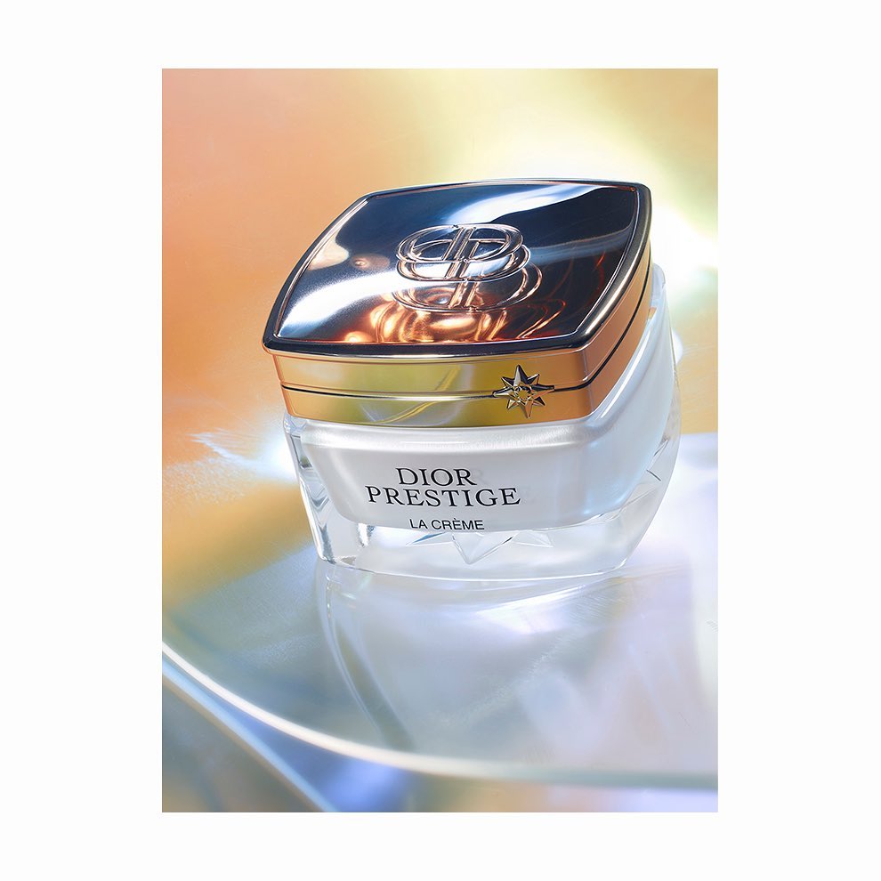 Dior Prestige La Crème jar with silver and gold lid on a reflective surface with a soft peach and yellow gradient background.
