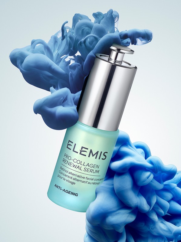 Elevated view of an Elemis Pro-Collagen Renewal Serum bottle, in focus, with a silver cap and teal label, surrounded by blue smoke.