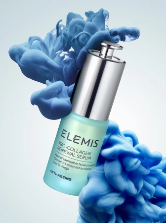 Elevated view of an Elemis Pro-Collagen Renewal Serum bottle, in focus, with a silver cap and teal label, surrounded by blue smoke.
