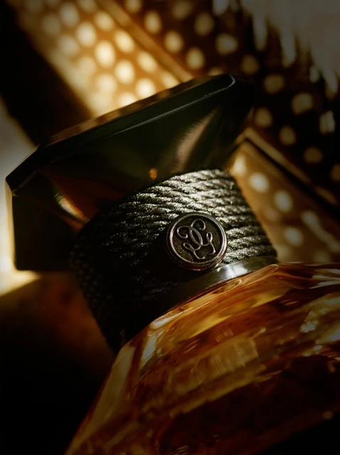 Close-up of a perfume bottle with a textured neck band and a geometric cap, set against a blurred background of spotted feathers.