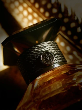 Close-up of a perfume bottle with a textured neck band and a geometric cap, set against a blurred background of spotted feathers.