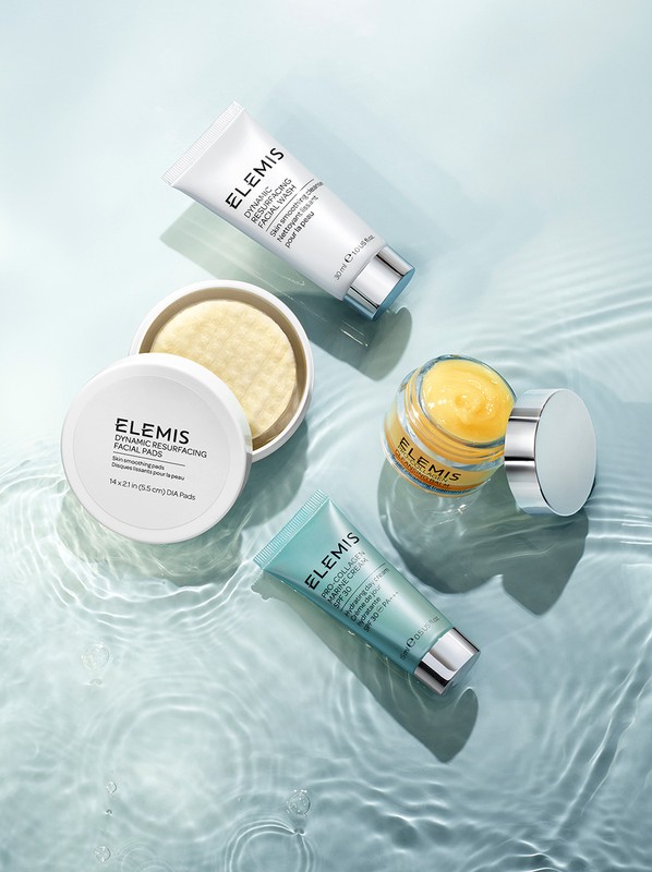 Overhead shot of assorted Elemis skincare products, including cream jars and tubes, arranged on a rippled glass surface for beauty advertising.