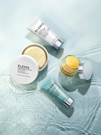 Overhead shot of assorted Elemis skincare products, including cream jars and tubes, arranged on a rippled glass surface for beauty advertising.
