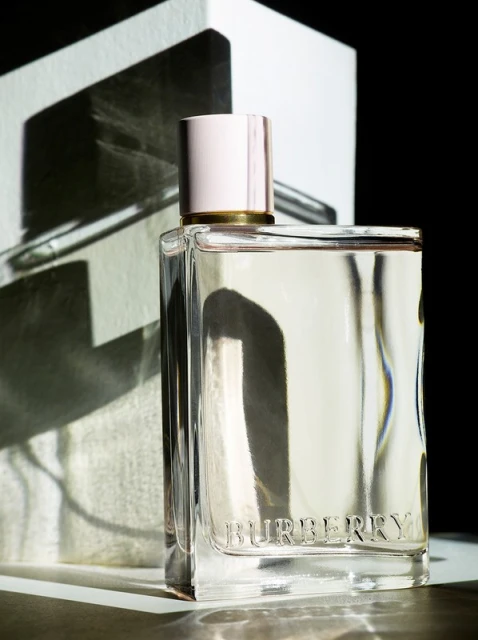 Elegant still life shot of Burberry perfume bottle with reflective box backdrop, set against a dark background in soft, diffused light.