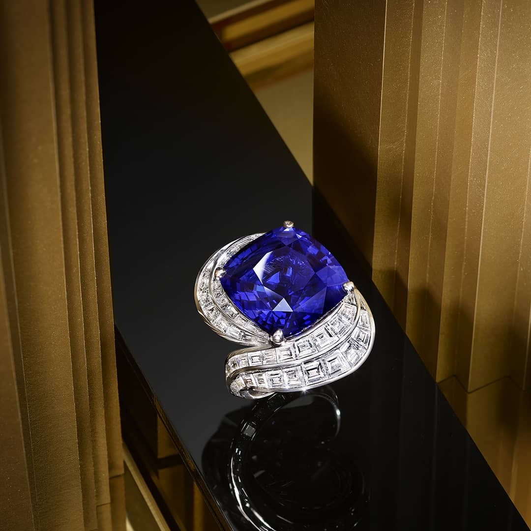 Cushion-cut tanzanite ring on a black glossy surface, surrounded by diamonds with a gold column background in a high-end jewelry context.