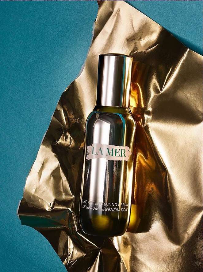 La Mer Regenerating Serum bottle displayed against a backdrop of blue paper and shiny gold material in a studio setting.