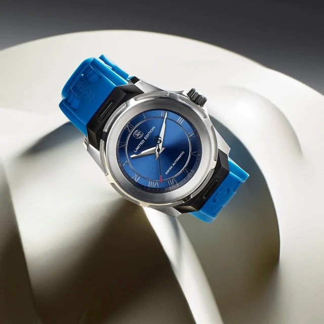 Silver and black wristwatch with a blue face and a blue rubber strap floating above a curved, white surface in a studio setting.