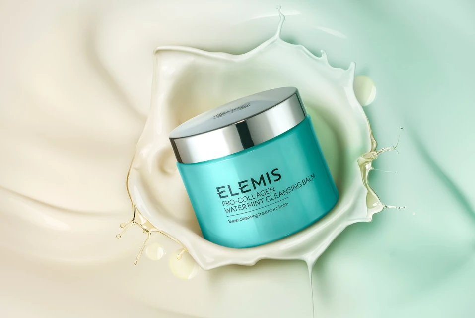 Turquoise jar of Elemis Pro-Collagen Water Mint Cleansing Balm surrounded by a swirl of white cream against a gradient backdrop.