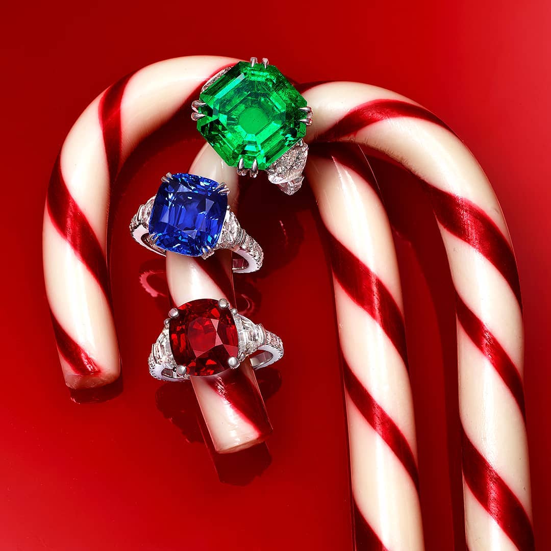 Three gemstone rings, one green, one blue, and one red, rest on a candy cane against a red background in a holiday jewelry advertisement.
