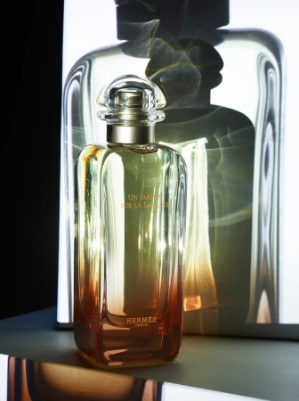 Elegant rectangular perfume bottle with clear cap, filled with a gradient of golden liquid, and the Hermes logo, stands in a studio setting.