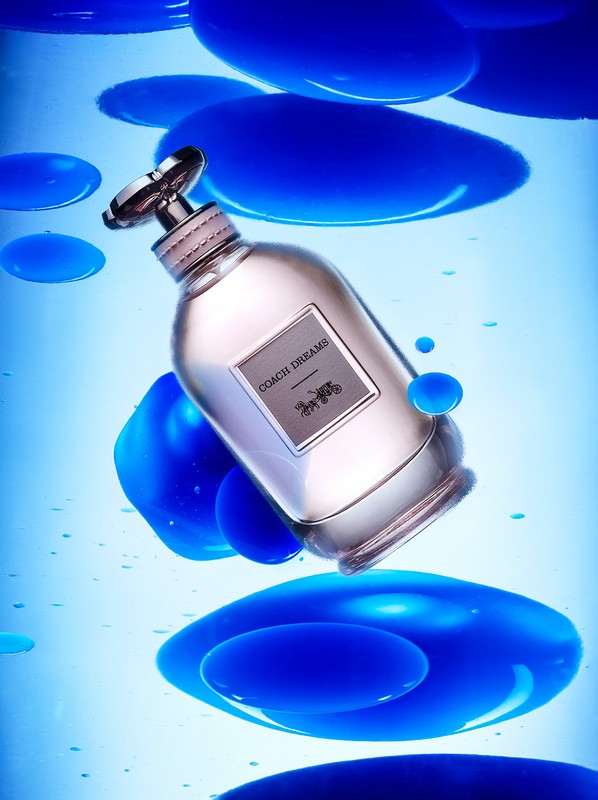 A silver and pale pink perfume bottle with the Coach Dreams label floats among blue gel shapes, all set against a pale blue background.