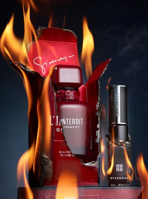 A perfume bottle and lipstick tube from Givenchy are dramatically presented, engulfed in flames against a dark background with subtle smoke.