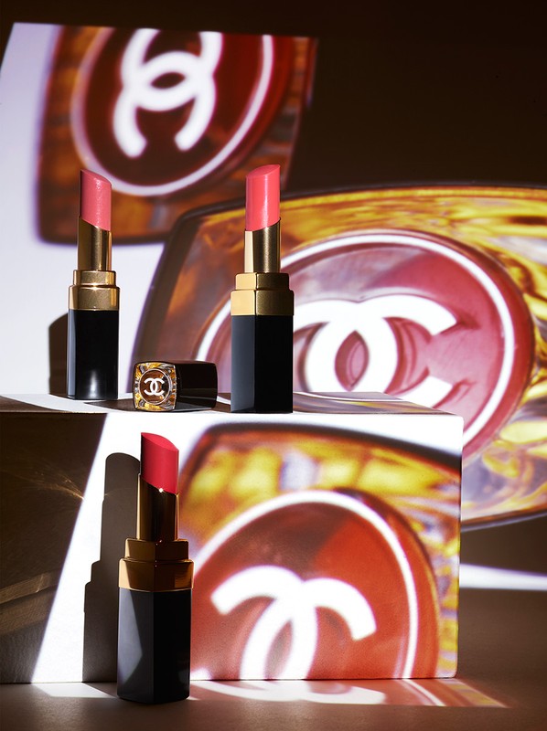 Three Chanel lipsticks are displayed against a backdrop featuring the Chanel logo, creating a luxurious and branded beauty product shot.