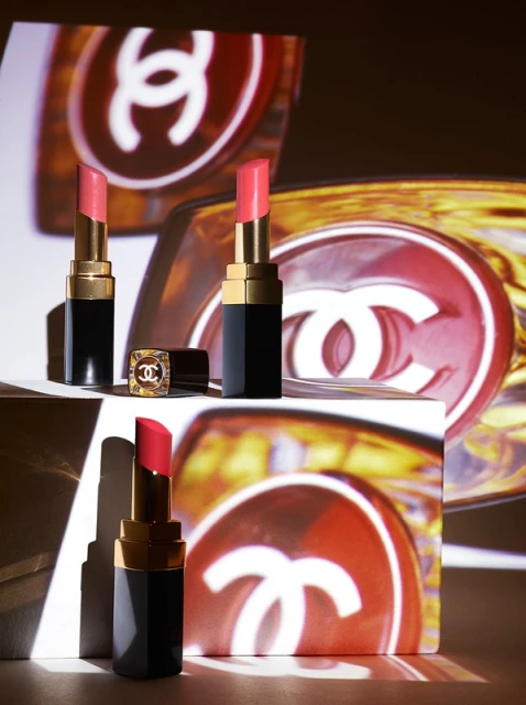 Three Chanel lipsticks are displayed against a backdrop featuring the Chanel logo, creating a luxurious and branded beauty product shot.