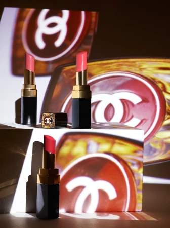 Three Chanel lipsticks are displayed against a backdrop featuring the Chanel logo, creating a luxurious and branded beauty product shot.